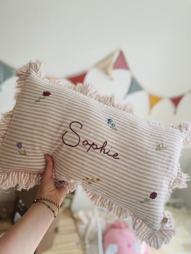 Personalised pink striped stonewashed linen cushion with ruffle trim, hand-painted ditsy florals and embroidered name, styled in a floral children’s bedroom.
