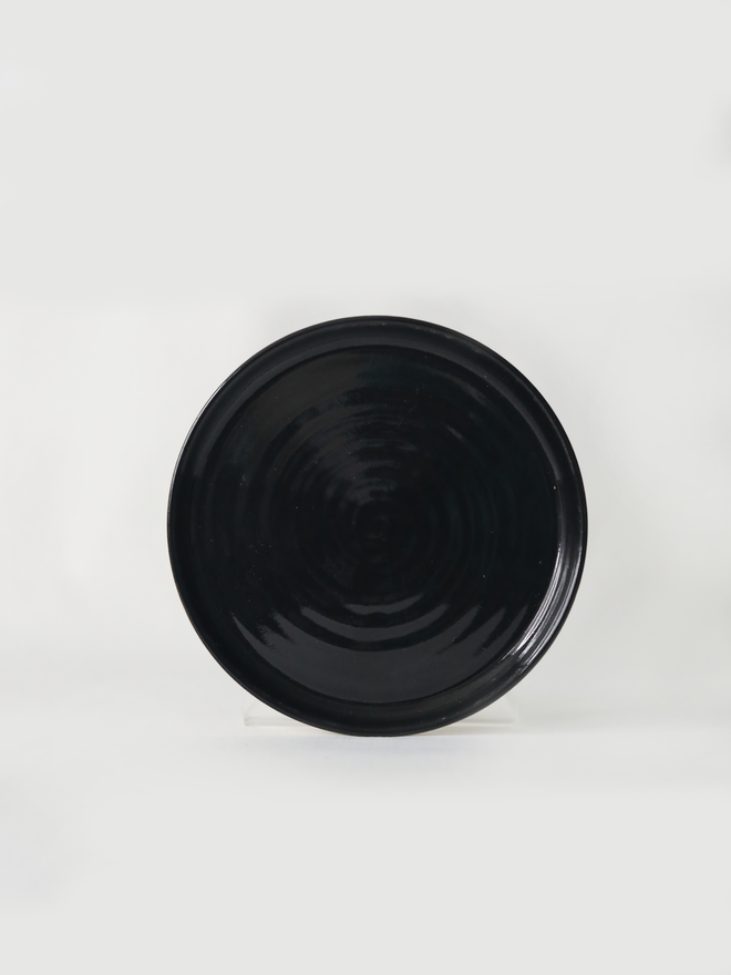 A handmade ceramic cake plate in jet black glaze, photographed from the front against a plain light grey background, showing a natural concentric swirl texture and a shallow raised rim