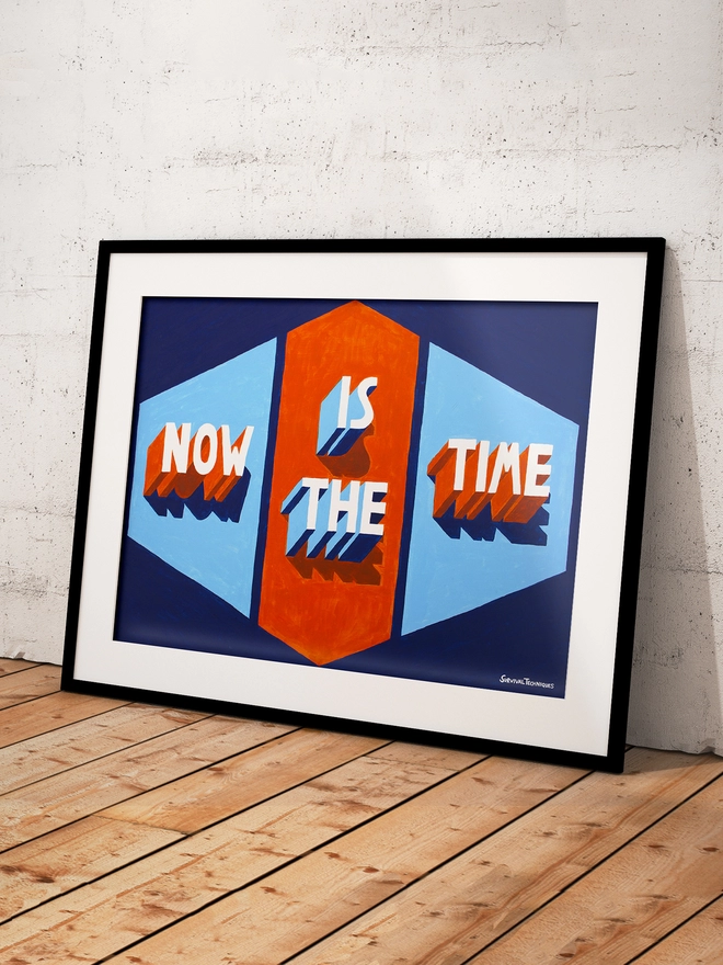 Framed art print of words Now Is The Time painted in 3d typography in orange and blue by artist Survival Techniques. Frame leans against wall on wooden floor.