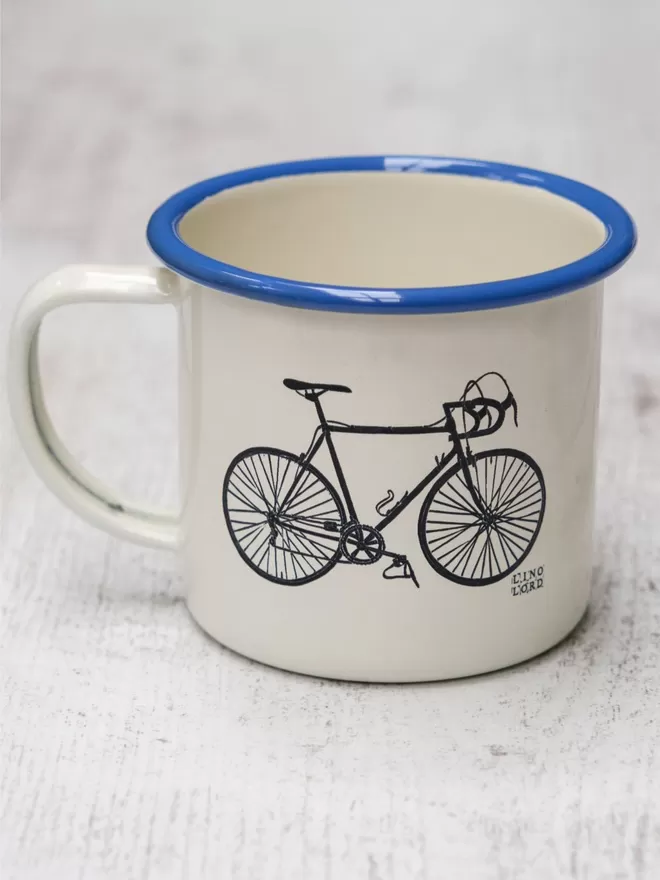 Picture of a Cream Enamel Mug with a Blue Rim with a Bicycle design etched onto it, taken from an original Lino Print