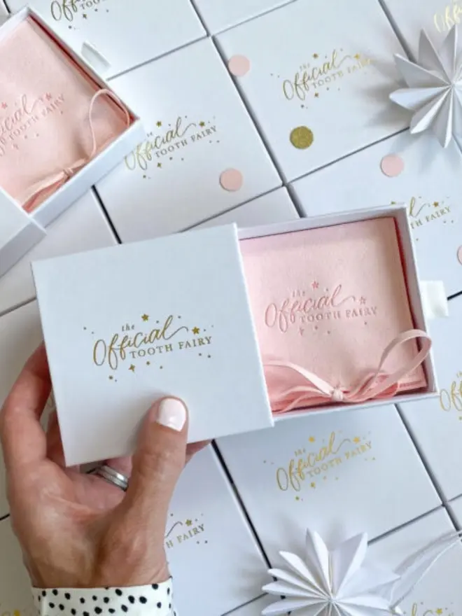 A hand holds an open gift box with a blush pink suede pouch tied with a ribbon. A second pink pouch in its box sits nearby on rows of gold foil boxes.
