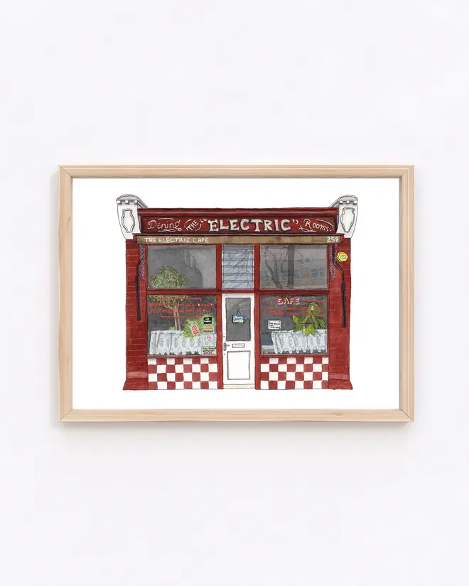 West Norwood The Electric Cafe Giclée Print Tulse Hill, a print of a cafe in a white frame sitting on a wooden surface against a white backdrop. 