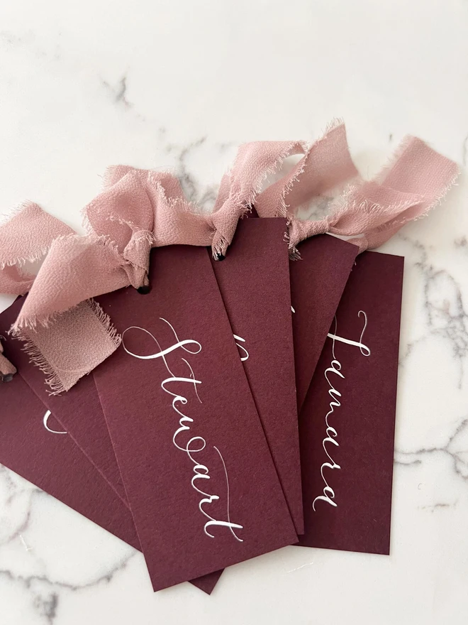 Personalised Calligraphy Wedding Burgundy Place Cards, burgundy place cards set together on a wooden table with chiffon plum ribbons. 