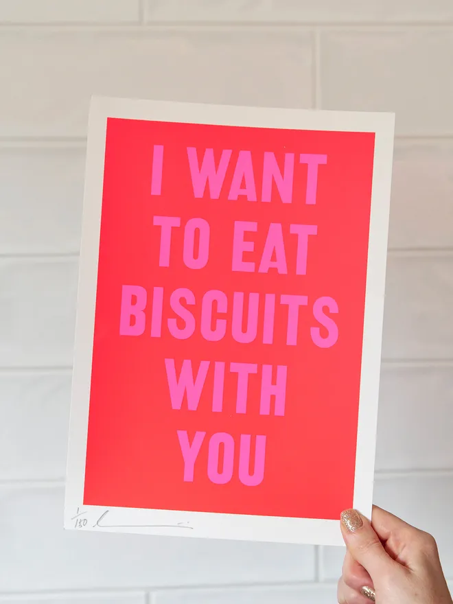 'I Want to Eat Biscuits with You' screen print in red with neon pink text, held up against a white tiled wall