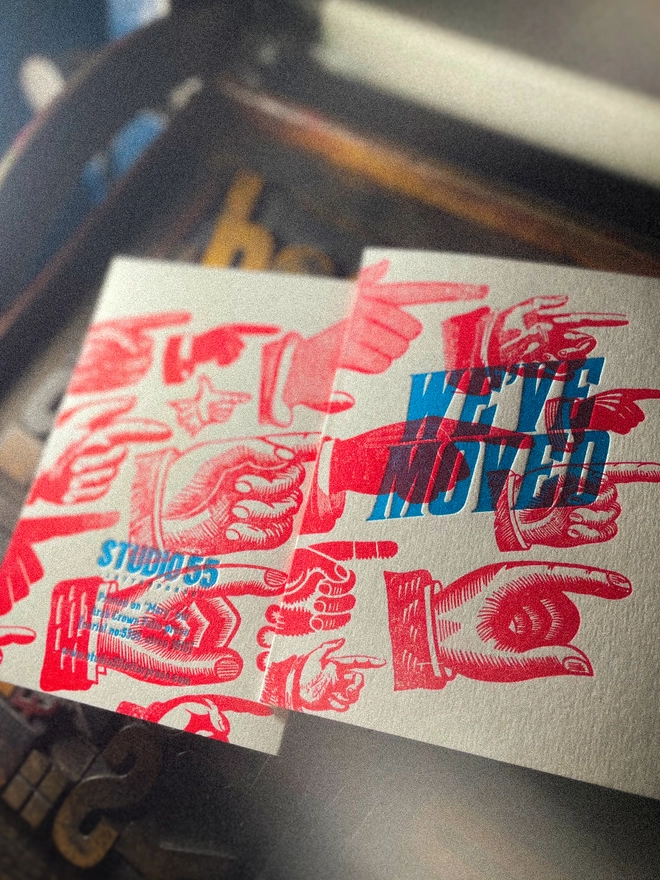 A double-sided, hand-printed letterpress artisan moving house announcement card with bold blue “We've Moved” typography and fluorescent red pointing hand illustrations (fists) on thick premium white card.