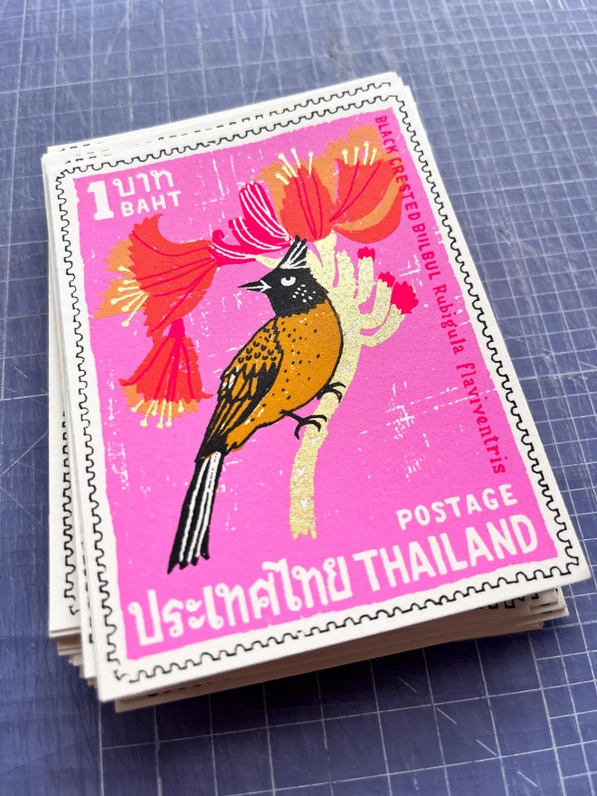 Bird Stamp Card Thailand Black Crested Bulbul, a white card with a colourful bird design in the centre on top of a stack of more cards on a studio surface. 