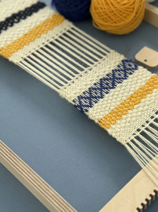 Wooden beige frame loom with strands of yarn and woven fabric in blue mustard and cream. 