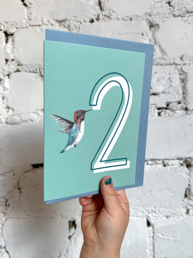 a pale teal card with a big white number 2 and an illustration of a hummingbird with a blue envelope