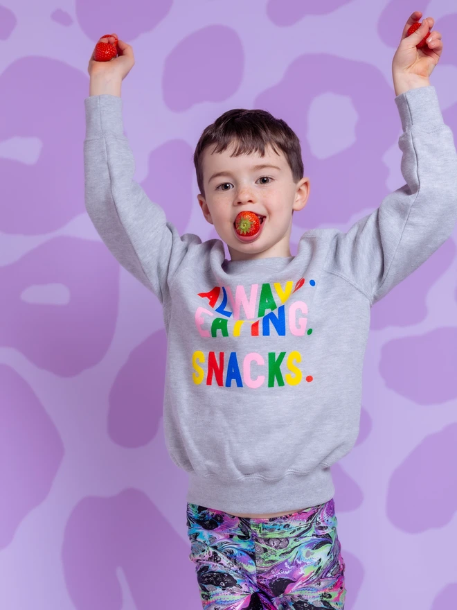 Child wearing a grey sweatshirt with Always Eating Snacks slogan