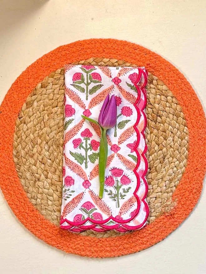 Close up of a round woven seagrass placemats with orange rings, shown on a light surface with a flowery napkin placed on top. 