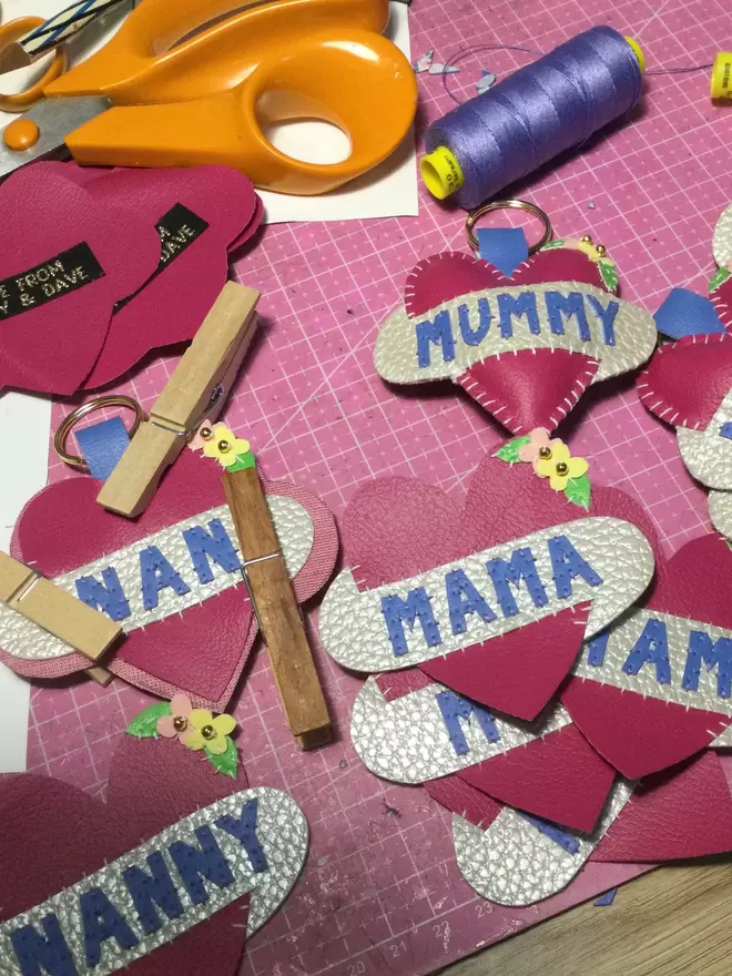 A work in progress - selection of pink leatherette hearts at various stages of being made, laying on a pink cutting mat. One says Nanny in blue lettering across a white scroll, others say NAN, MAMA, MOTHER, MUMMY and MUM