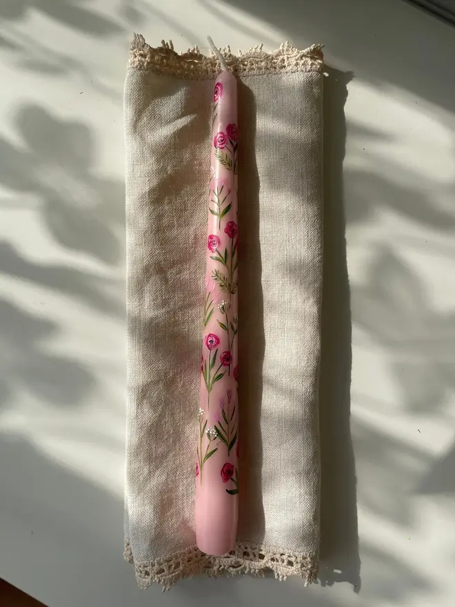 Pink Blooms Tapered Dinner Candle, a pink candle lying on a napkin on a table surface. The floral design wraps around the stalk. 