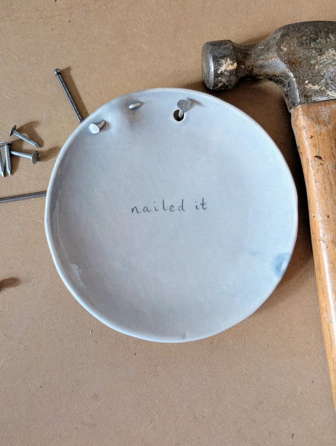 congratulations wall art grey plate with grey glaze and sculpted nail through the rim an the pun nailed it hand painted next to hammer and nails 