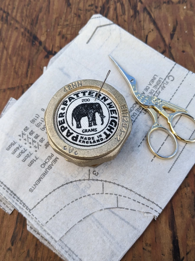 Black elephant brass pattern & paper weight with needle lying on a sewing pattern next to scissors