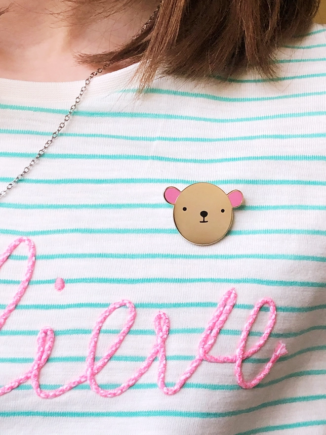 A gold bear pin with pink ears is pinned on a white and green striped top.