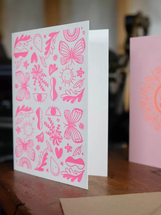 Pink letterpress printed shapes including a butterfly, sun, eye, heart and flowers printed on an A6 portrait white card. Standing on a shelf. 