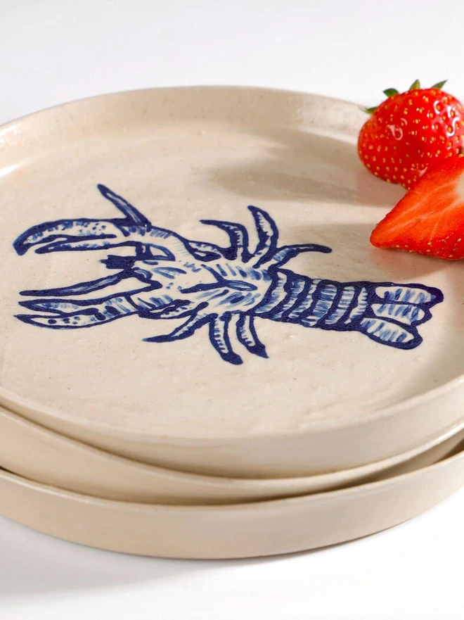 Handmade Ceramic Side Plate With Blue Lobster Design, a side plate with a blue lobster design sitting on a white table surface accompanied by a raspberry. 