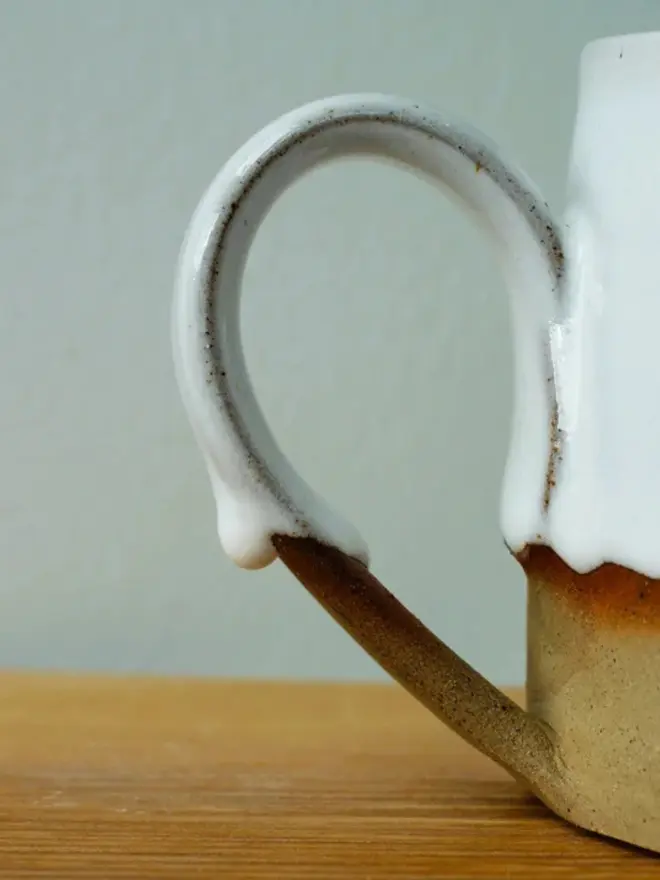 Handmade ceramic mug with a glossy white upper glaze and a natural toasted clay base, against a soft green wall background.