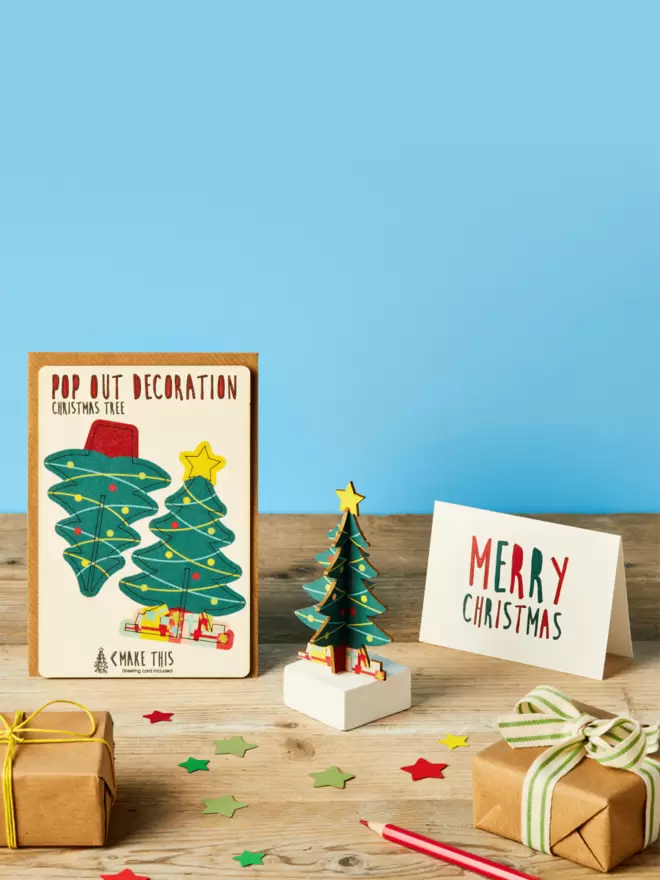Pop out Christmas tree decoration and Merry Christmas card and brown kraft envelope on top of a wooden desk in front of a sky blue coloured background