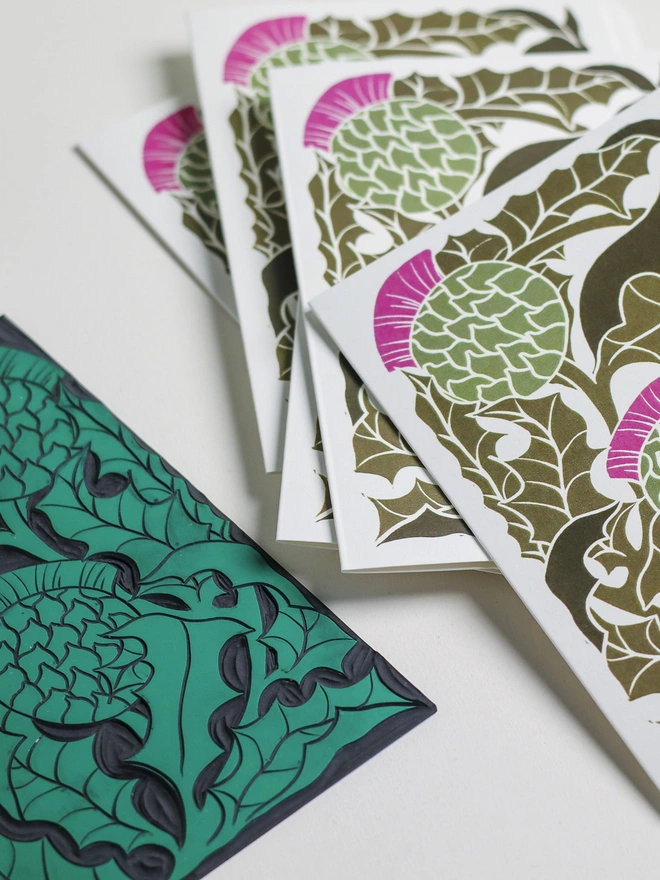 Hand-carved lino block displayed with finished Scottish Thistle notecard from set of 8. Traditional relief printmaking block showing carved botanical design alongside contemporary printed card. Authentic linocut process, Norfolk countryside studio.