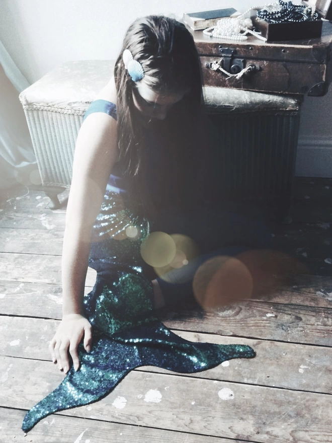 a Mermaid sat on wooden floor boards with her knees curled in front, wearing a peacock green sequin mermaid tail, a leotard and shells in her hair.  there is a suitcase filled with pearls positioned behind her 
