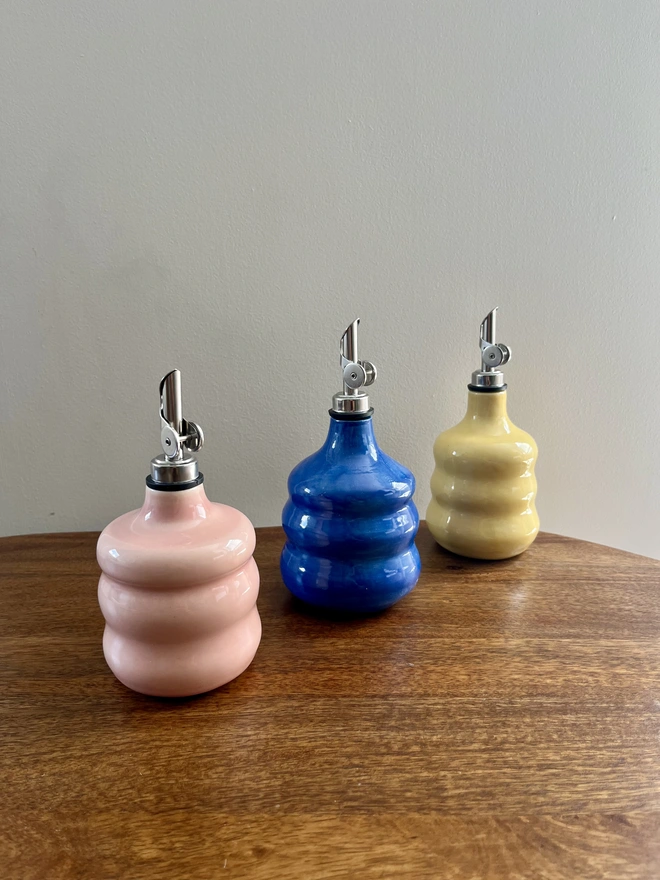 a collection of 3 colourful chubby oil bottles with wavy sides on a wooden table. blush pink, cobalt blue and butter yellow