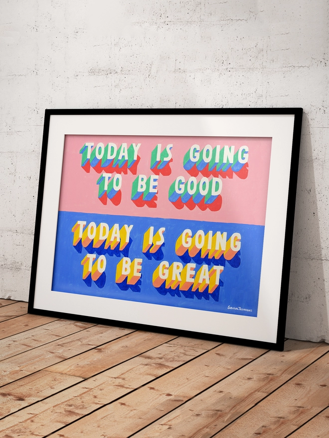 Framed art print of words Today Is Going To Be Good Today Is Going To Be Great painted in 3d typography in pink, green, yellow and blue by artist Survival Techniques. Frame leans against wall on wooden floor.