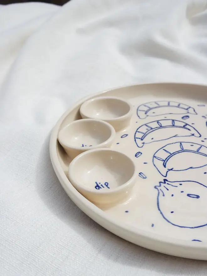Dumplings Dippy Dinner Plate, a dipping plate sitting on a white bed cloth. It is covered in blue outlines for dumplings. 