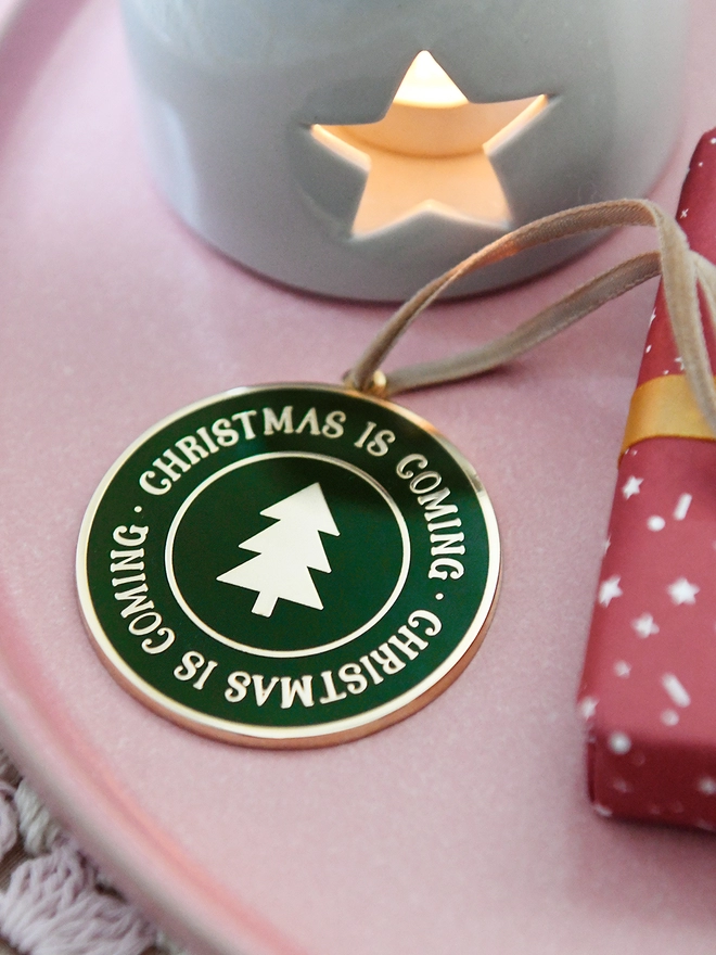 A deep green and gold enamel Christmas decoration, with the words Christmas Is Coming surrounding a gold Christmas tree, lays on a pink plate.