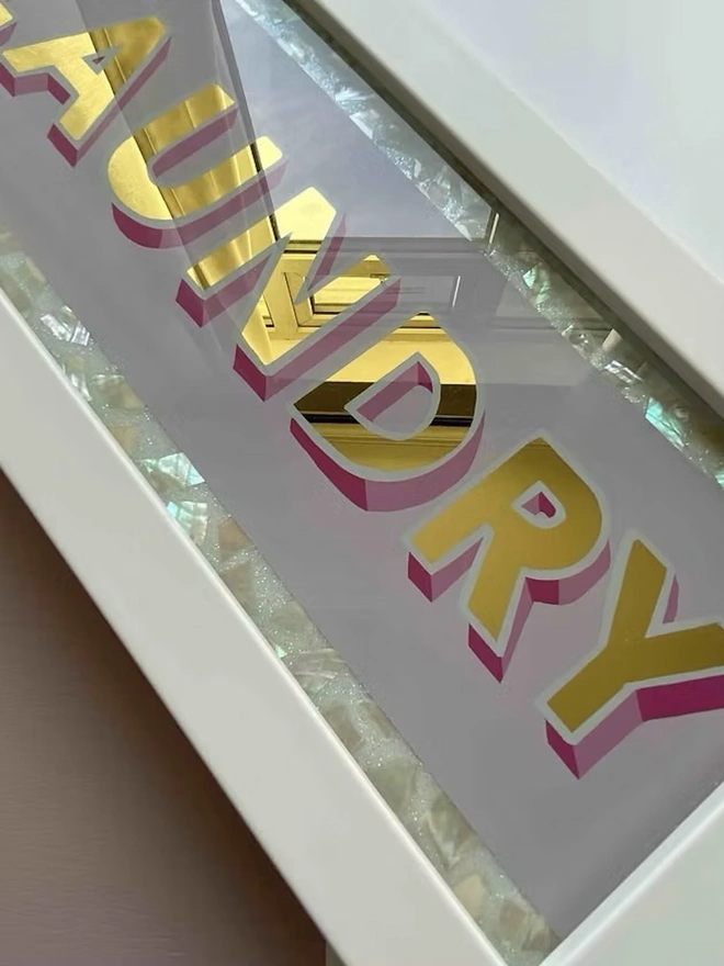 'Laundry' Glass And Gold Leaf Sign, a gold and glass sign sitting on a plain surface in a white frame. 