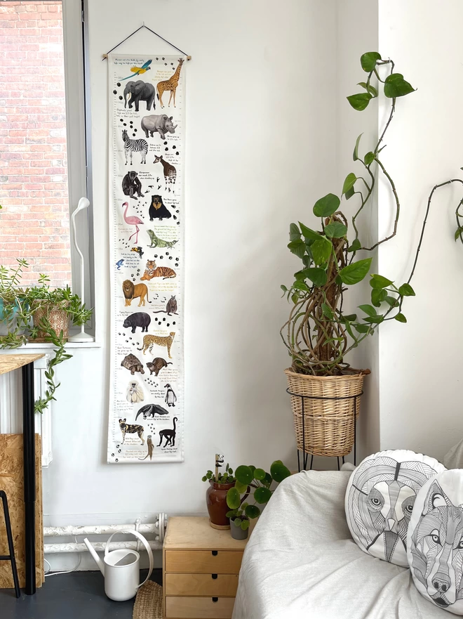 A photo of the full fabric height with lots of illustrated animals on it, on the wall in a room next to a window and a sofa