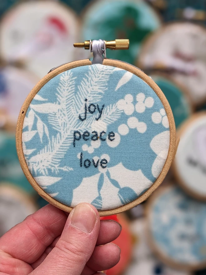 A hand embroidered hoop on Christmas fabric reading joy peace love.