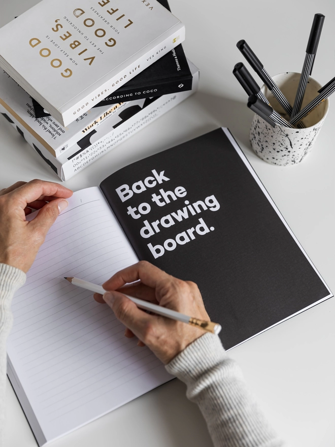 Lady writing on a lined page of the planner opposite a quote page reading "Back to the drawing board". 