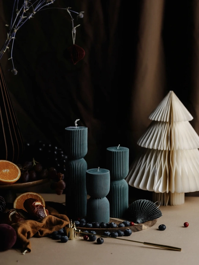 Forest Green Christmas Cracker Candles (Set Of 3). Three dark teal ribbed pillar candles with cinched waists, on a wooden board with blueberries and a paper fan beside a pleated paper tree, against a moody draped backdrop. The deep jewel tone and sculptural shape make a striking festive centrepiece.