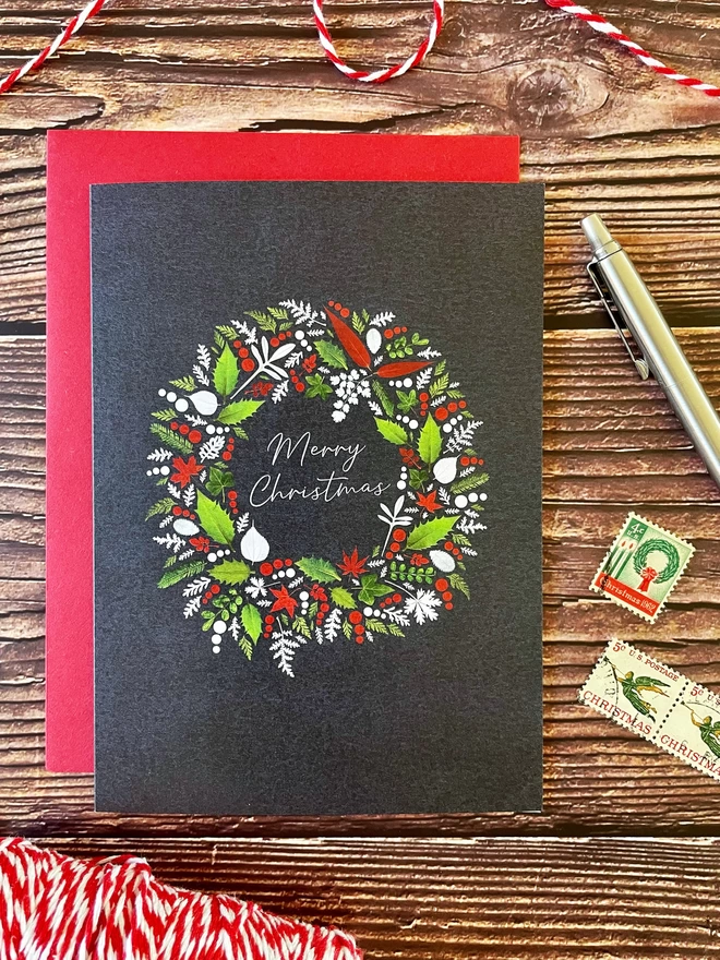 Digitally Printed Christmas Card with pressed Winter Foliage Design and Red Envelope on Wooden Background with Vintage Stamps, “Red and White Twine Spool and Silver Ballpoint Pen