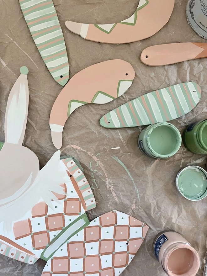 Large Hope Hare Wall Puppet. the individual painted wooden pieces of the large hope hare wall puppet are laid out on brown paper before assembly, showing the arms, legs and ears in soft pink and green tones with dotted and striped detailing. small pots of sage green and blush pink paint sit among the pieces, along with a partially assembled section of the hare's body and diamond-patterned skirt. the flat-lay view highlights the handcrafted, hand-painted nature of the puppet.