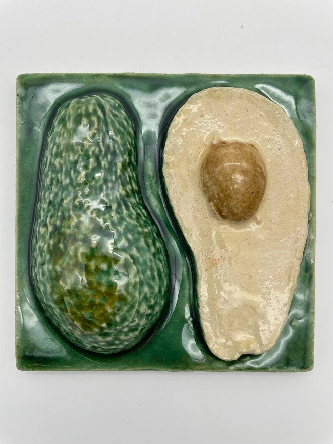 Handmade ceramic tile taken from a plaster cast of a real avocado, cut in half to show one half fleshy side up and one half skin side up. Very realistic, three-dimensional, with lush coloured glazes.