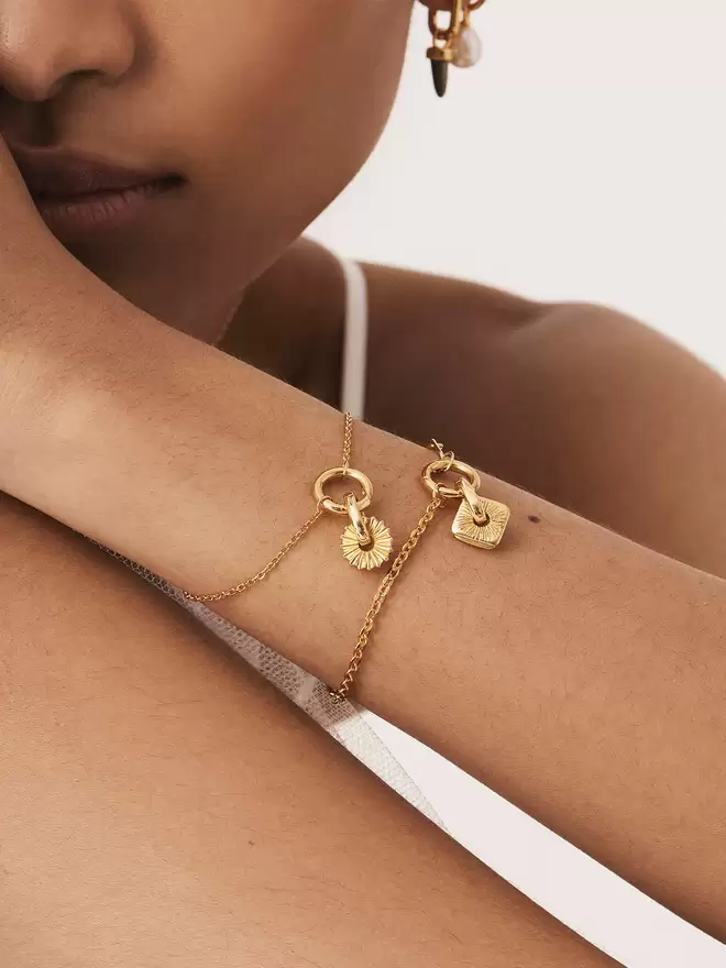 woman wearing two gold bracelets styled with a gold helios sun disc charm and a Doradus star square disc charm
