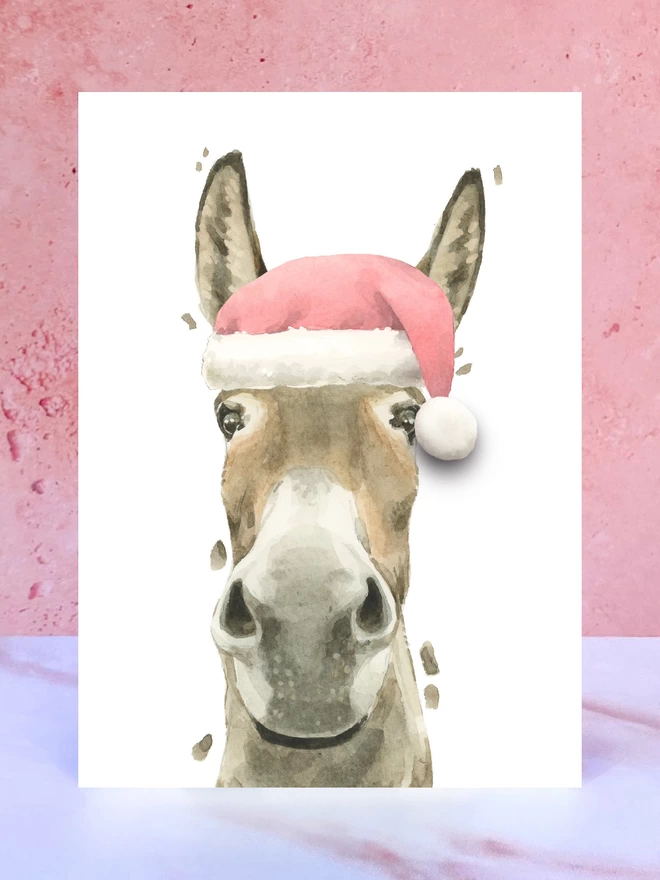 A Christmas card featuring a hand painted design of a donkey, stood upright on a marble surface. 