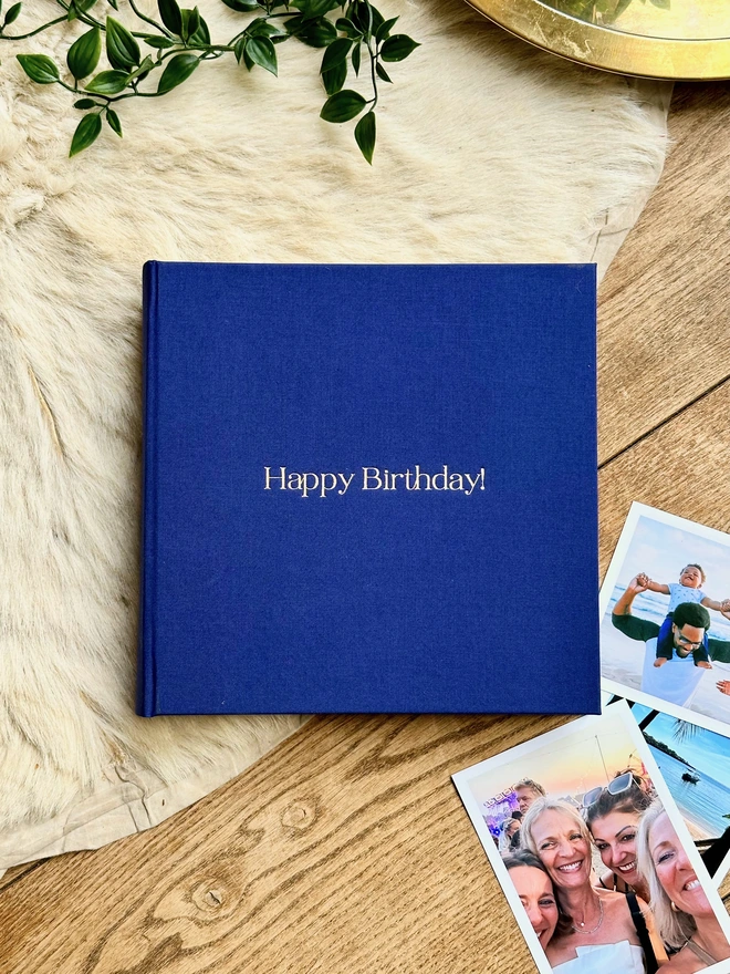 a blue linen photo album with the words Happpy Birthday printed on the front has been placed on a sheepskin rug. There are lots of photos surrounding the album of friends and family.