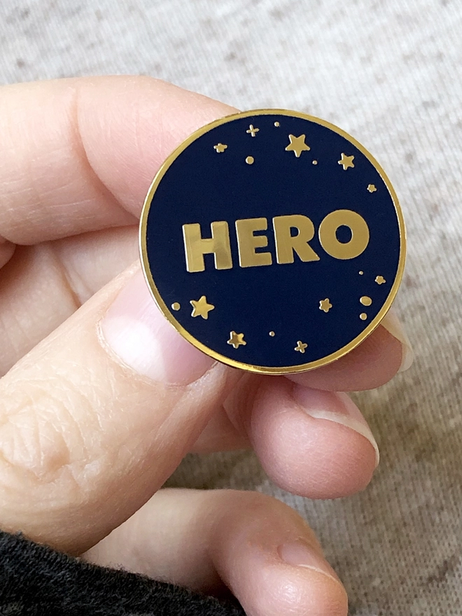 A navy blue and gold enamel pin badge with a starry design and the word "Hero" is being held.