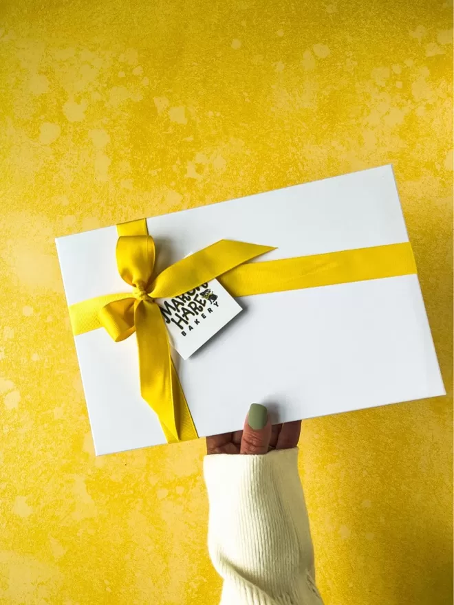 a female hand  with sage green nails holding up a white gift box with a thick yellow ribbon tied around it , against a bright yellow background