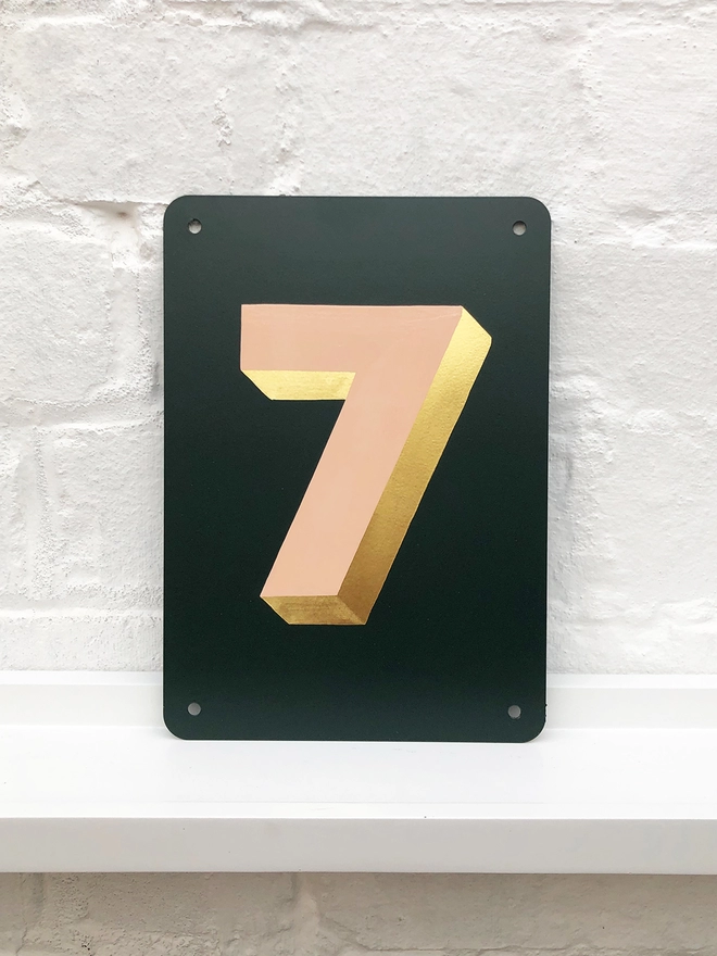 Hand painted peach and gold leaf house number 7 on an anthracite grey metal plaque against a white brick wall. 