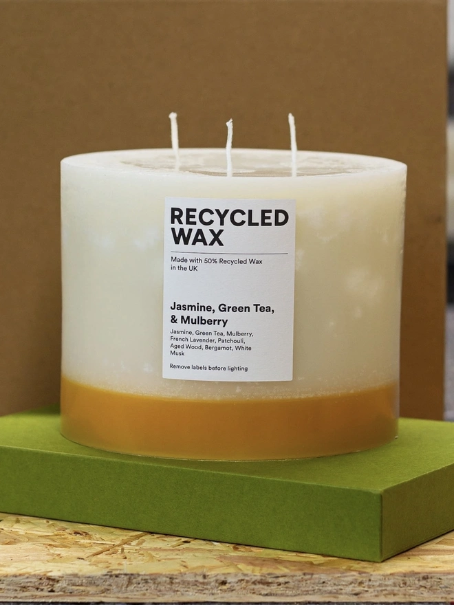 Jasmine, Green Tea & Mulberry Candle (Set Of 1 Or 3 Wicks).  the jasmine, green tea and mulberry candle stands on a green box against a brown background, showing its three wicks and cream wax with a golden yellow base. a white label reads "recycled wax, made with 50% recycled wax in the uk", followed by the scent name and note list of jasmine, green tea, mulberry, french lavender, patchouli, aged wood, bergamot and white musk. the candle's two-tone colouring and visible drip texture give it a rustic, hand-poured look.