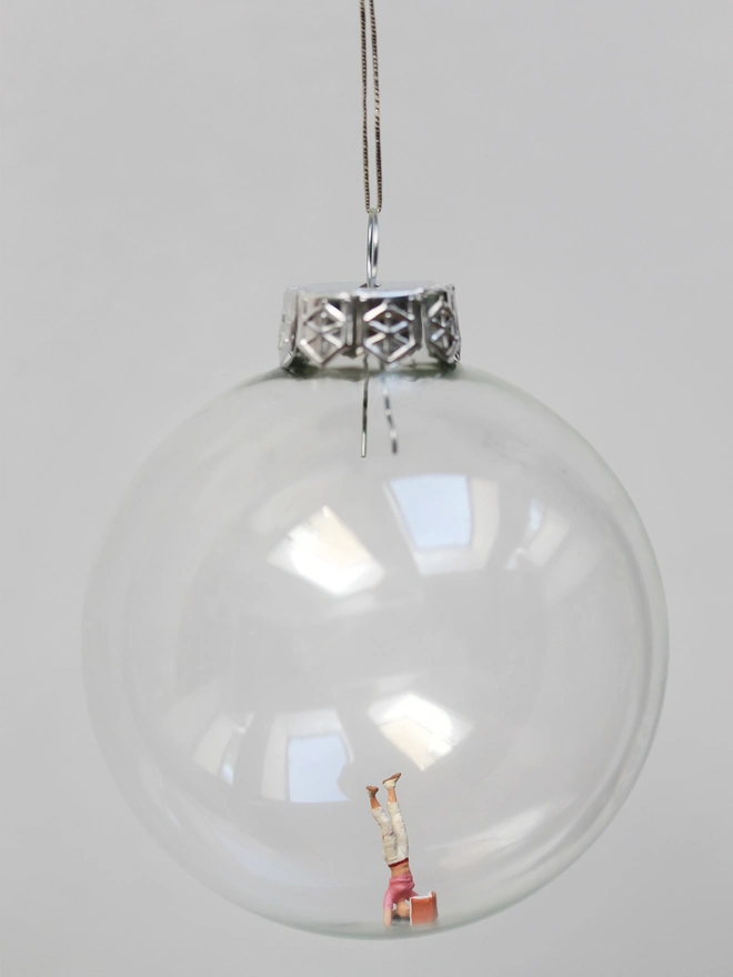 Miniature figure of a woman reading while doing a headstand, in a glass Christmas bauble 