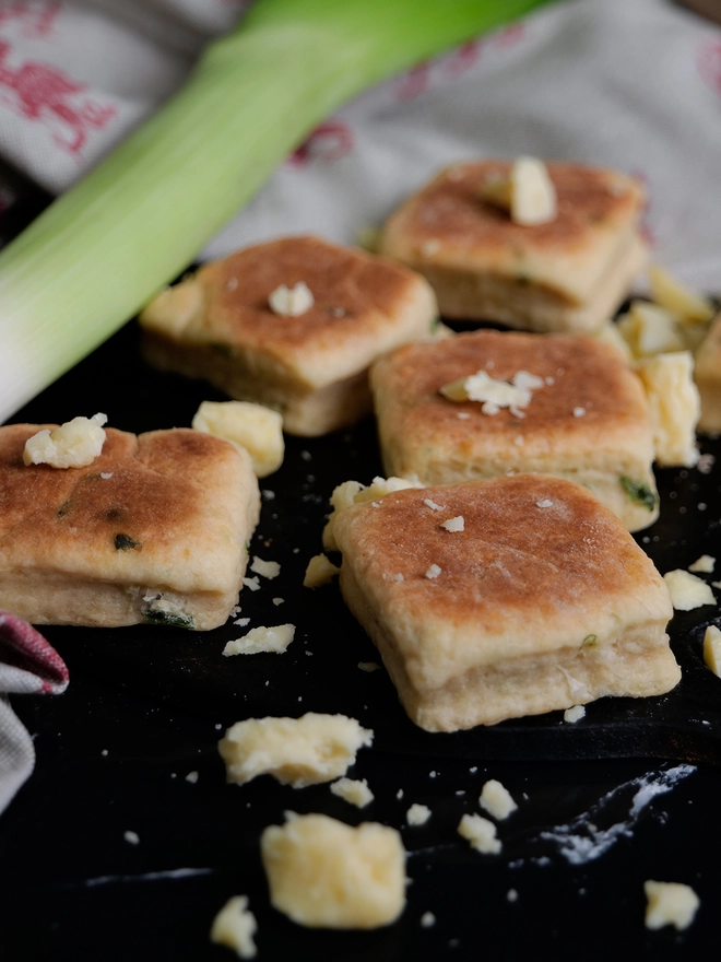 Cheese and Leek Welsh Cakes on a griddle with broken cheese and a large leek