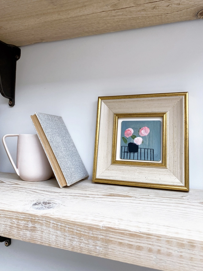 pale wooden shelf with framed picture of three painterly pink flowers on dusty blue background 