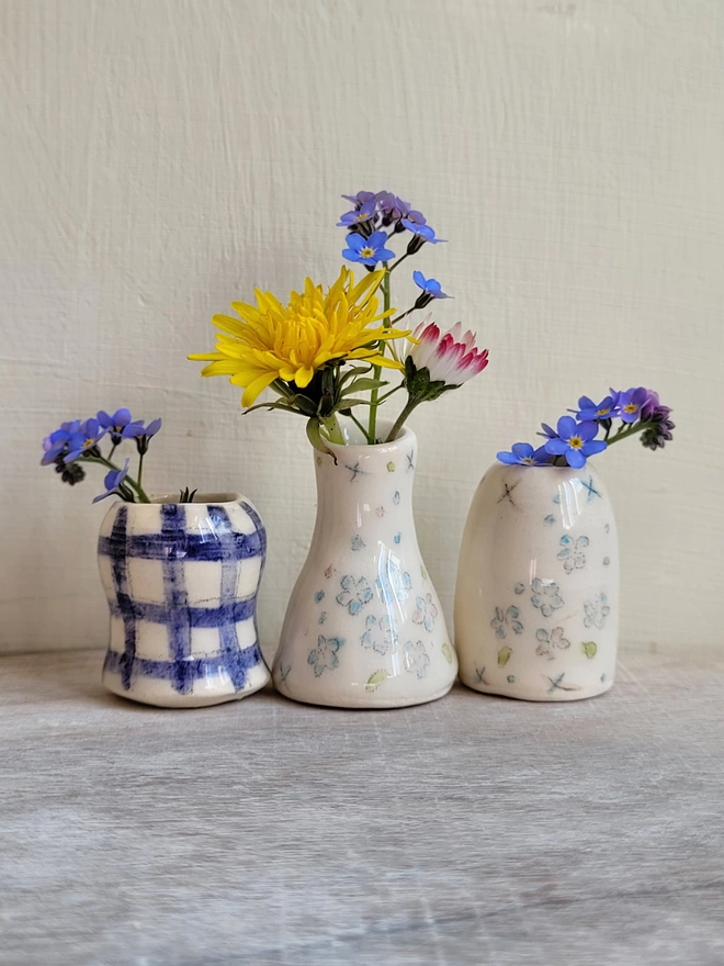 three miniature pottery vases with hand painted forget nots and blue gingham in a row on a wooden surface with daisies, forget me not flowers and a dandilion in