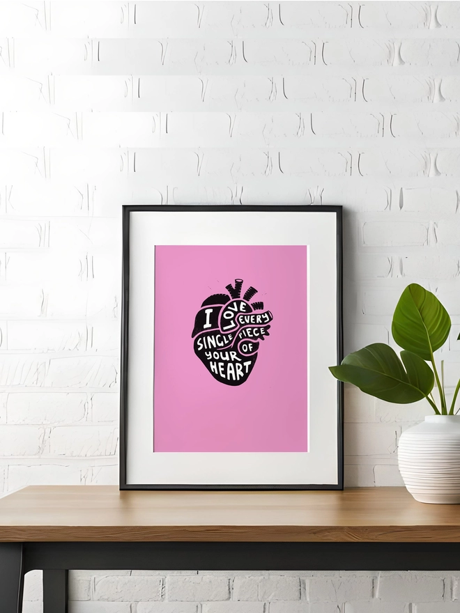 A pink Woodism print in a black frame. The print is pink with a linoprint design of a black anatomical heart in the centre. In white hand carved type it reads 'I Love Every Single Piece of Your Heart.'  