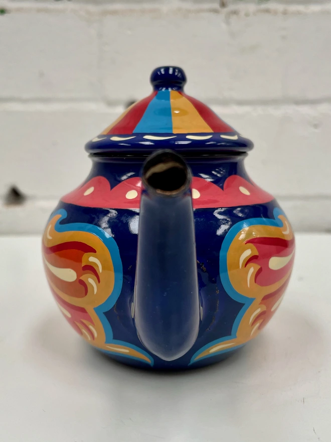 Small blue vintage enamel tea pot, hand painted with a fairground style design in pink, orange, and red. Teapot has a hinged lid.